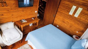 A wooden room with a bed, a chair, a table with books, and a TV mounted on the wall.