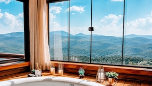 A room with a large window, a wooden floor, a white bathtub, and a view of mountains.
