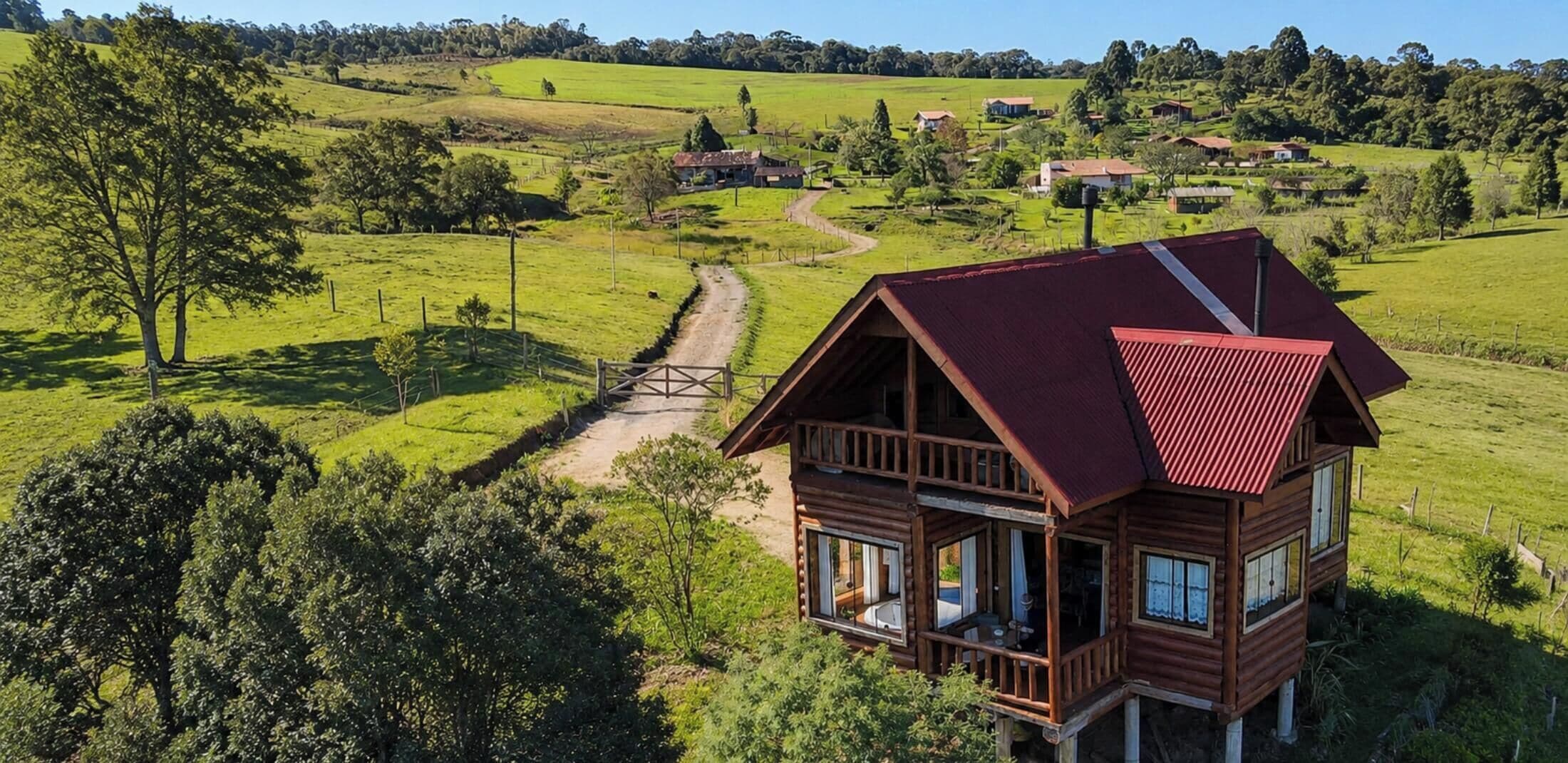 Uma cabana de madeira com telhado vermelho, rodeada por campos verdes exuberantes e árvores, com um caminho de terra que a leva.