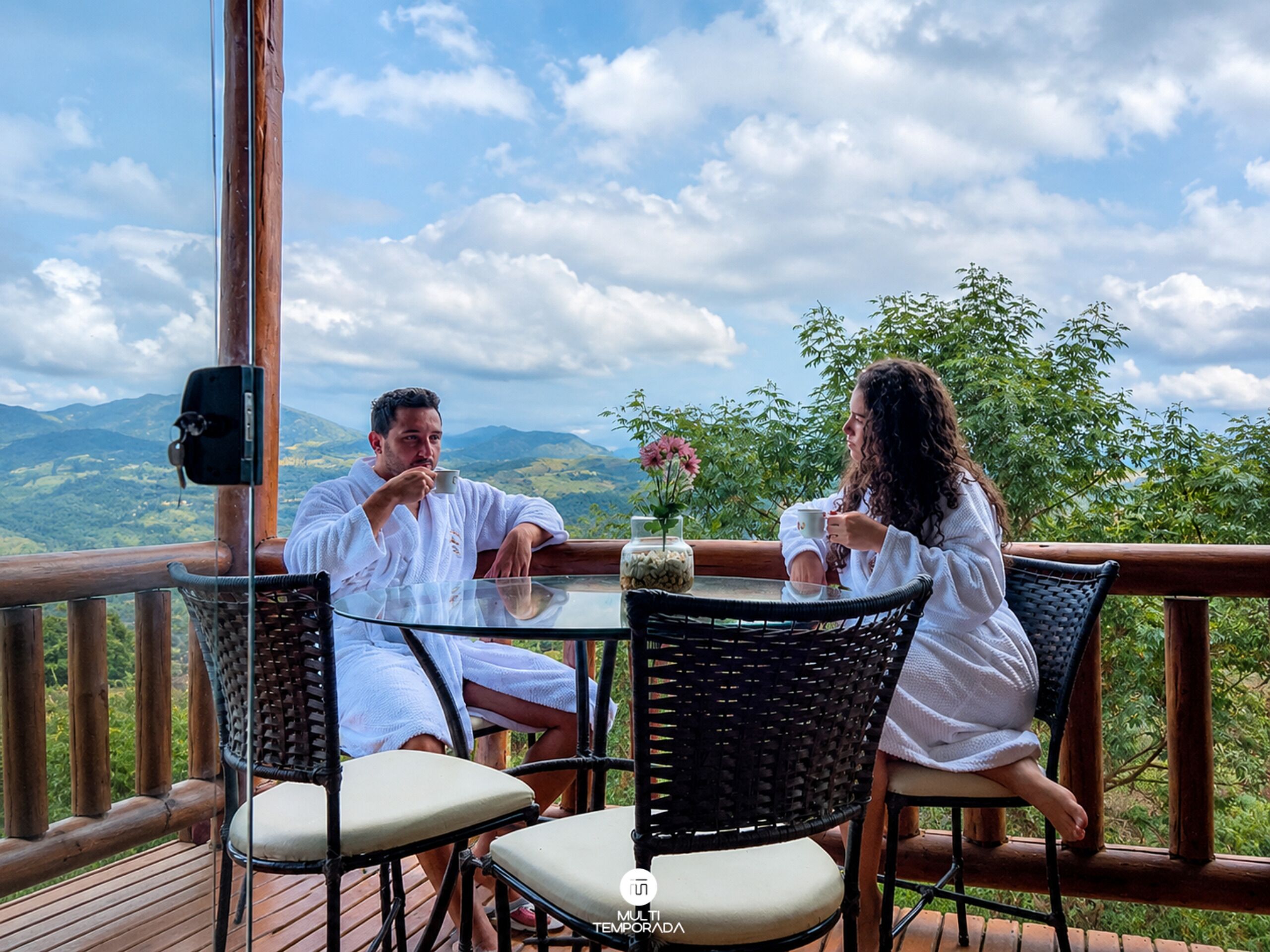 A couple seated at a table with a scenic mountain view, wooden deck, and wicker chairs.