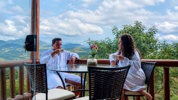 A couple seated at a table with a scenic mountain view, wooden deck, and wicker chairs.