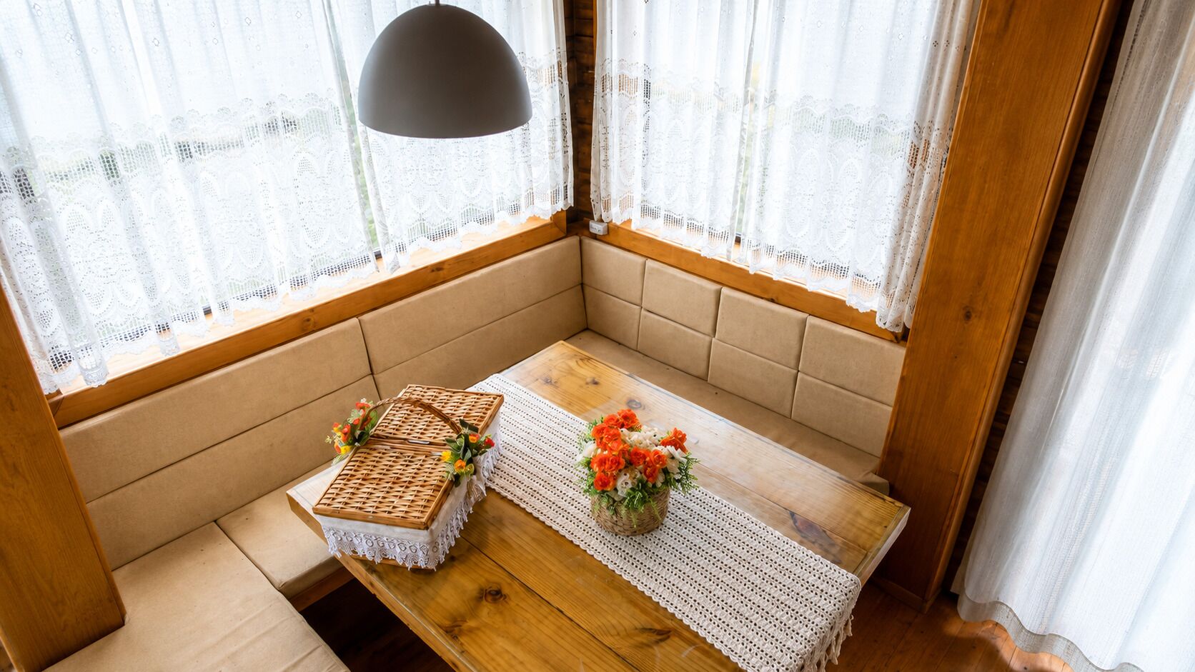 A corner seating area with a wooden table, lace table runner, wicker baskets, and a floral arrangement.