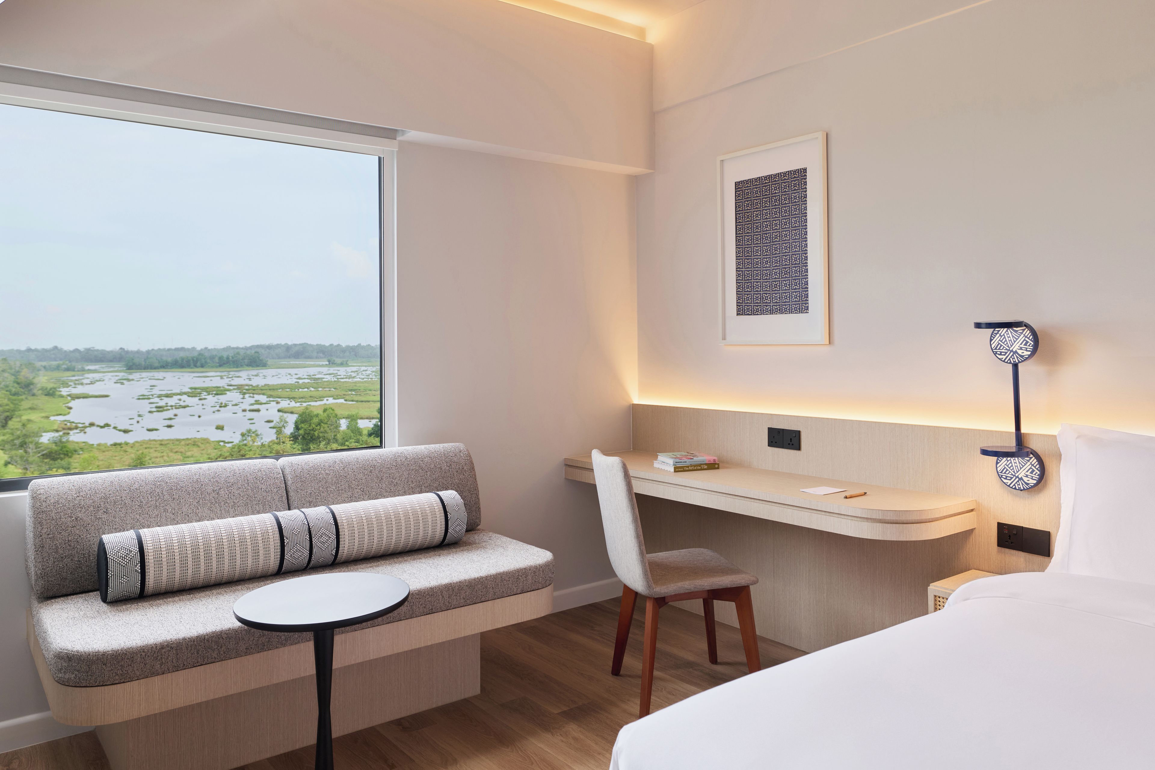 Une chambre d’hôtel avec une grande fenêtre, un bureau, une chaise, un lit et une lampe fixée au mur.
