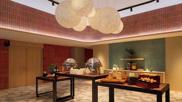 A hotel lobby with a high ceiling, red brick walls, and a modern chandelier. There are tables with breakfast items and a reception desk.
