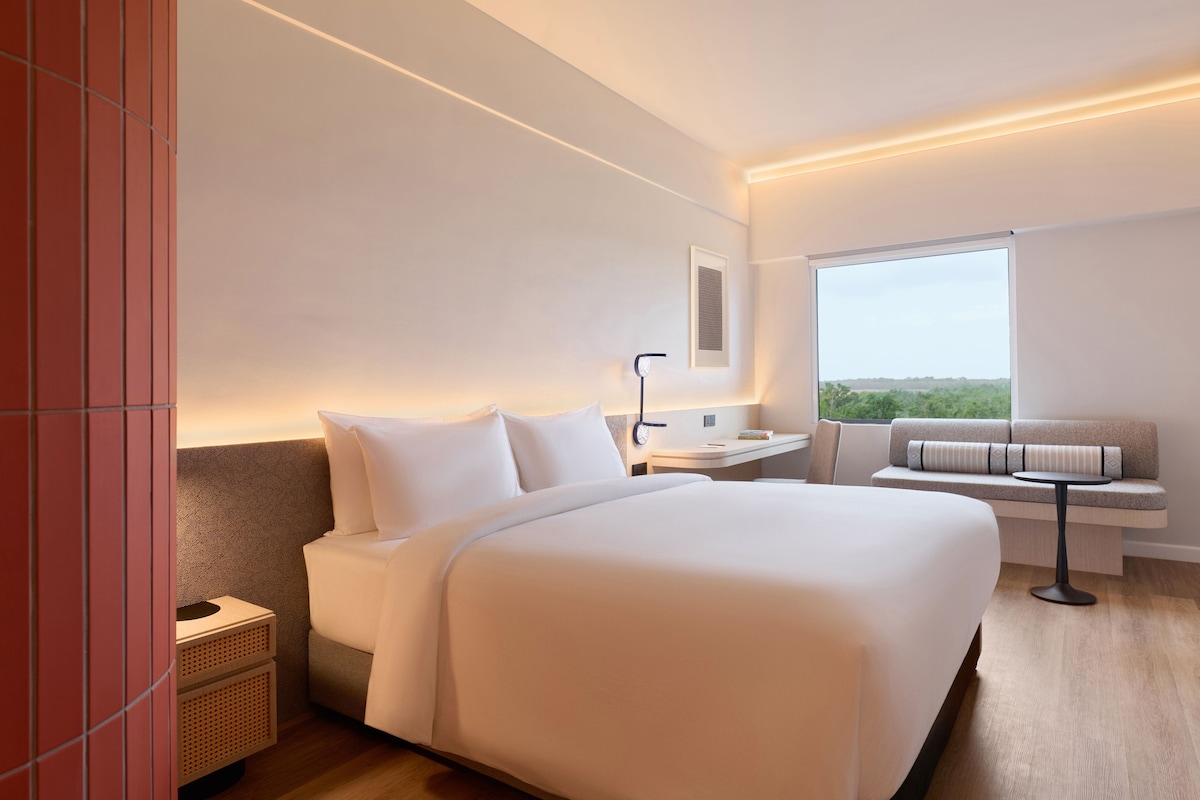 A modern hotel room with a large bed, a bedside table, a reading lamp, a sofa with a view, and a window.