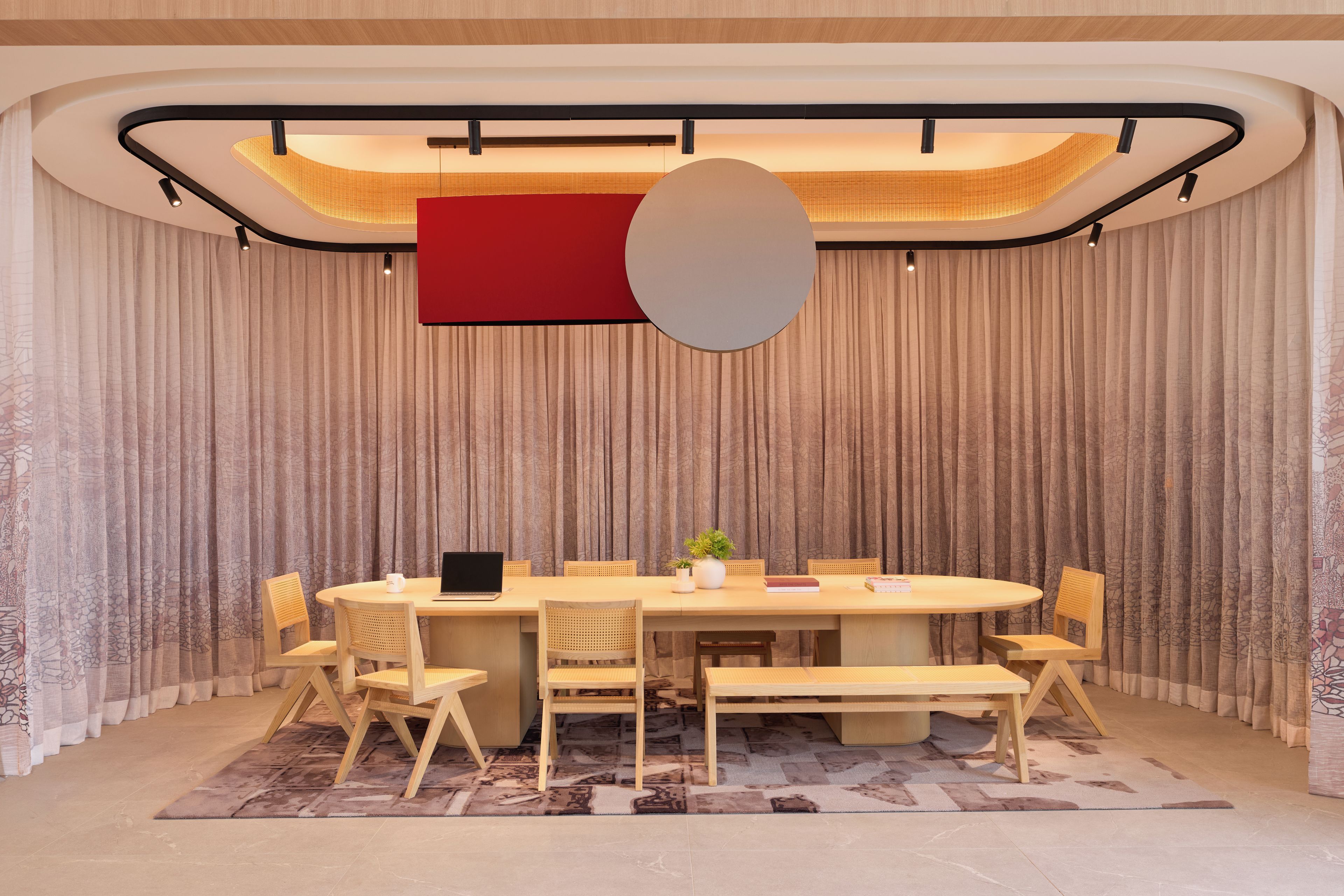 A conference room with a round table, wooden chairs, a laptop, a plant, and a red accent wall.