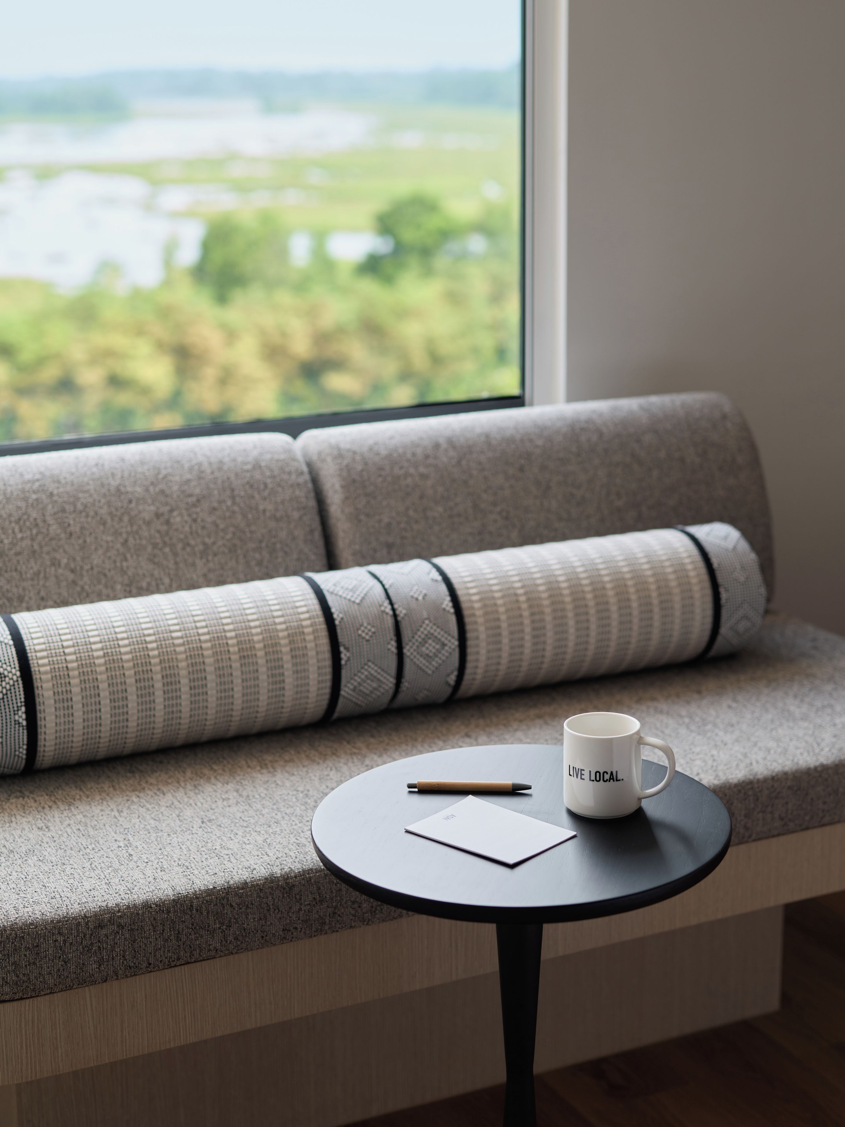 A sofa with a patterned cushion, a small round table with a mug and a pen, and a view of greenery through a window.