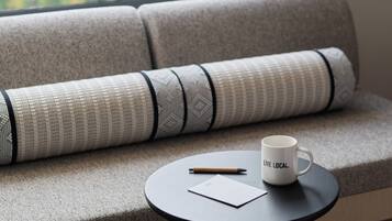 A sofa with a patterned cushion, a small round table with a mug and a pen, and a view of greenery through a window.