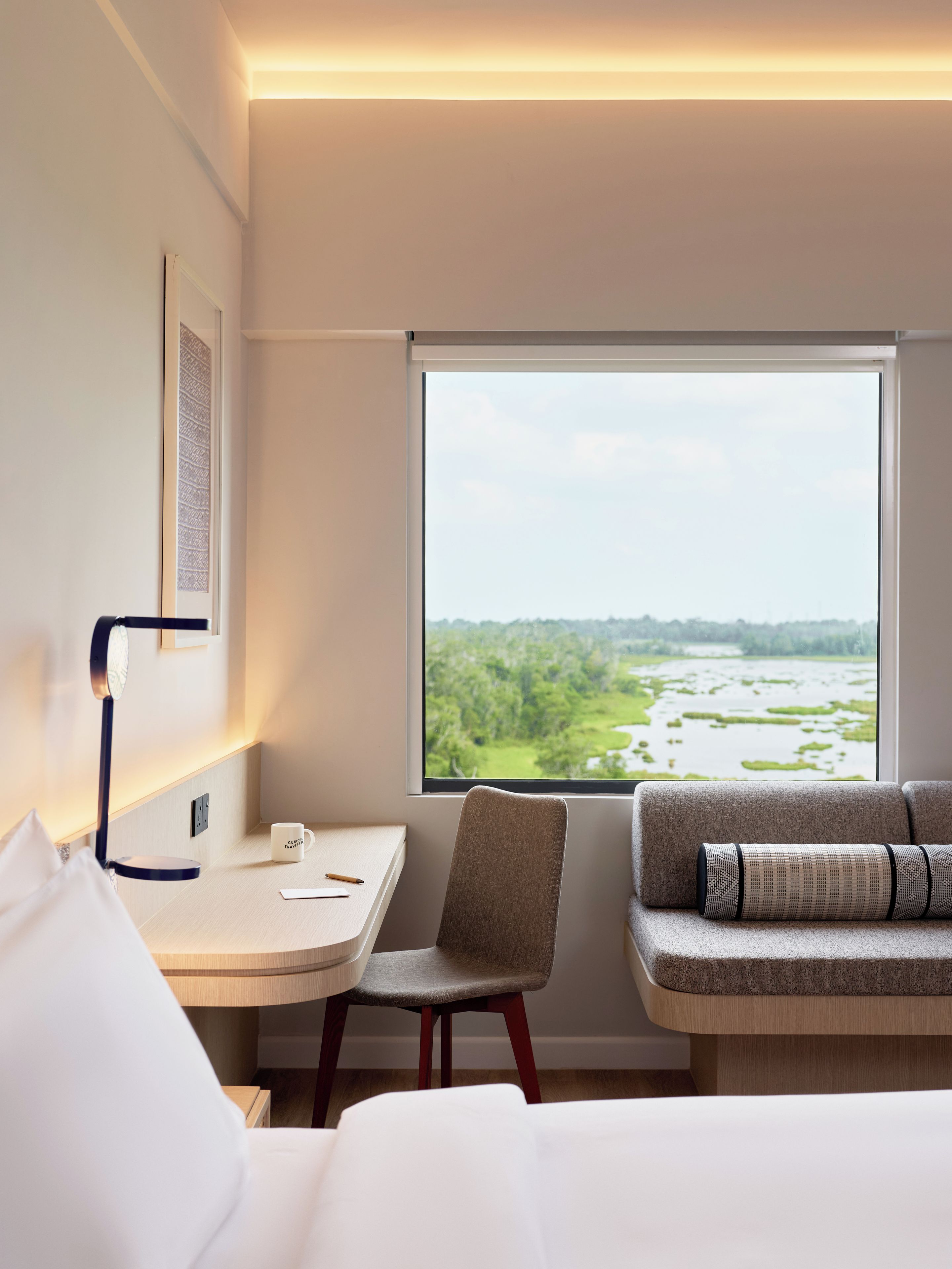 Une chambre d’hôtel comprenant un lit, un bureau avec une chaise, une fenêtre donnant sur un espace verdoyant et une lampe fixée au mur.