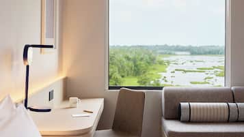 Une chambre d’hôtel comprenant un lit, un bureau avec une chaise, une fenêtre donnant sur un espace verdoyant et une lampe fixée au mur.