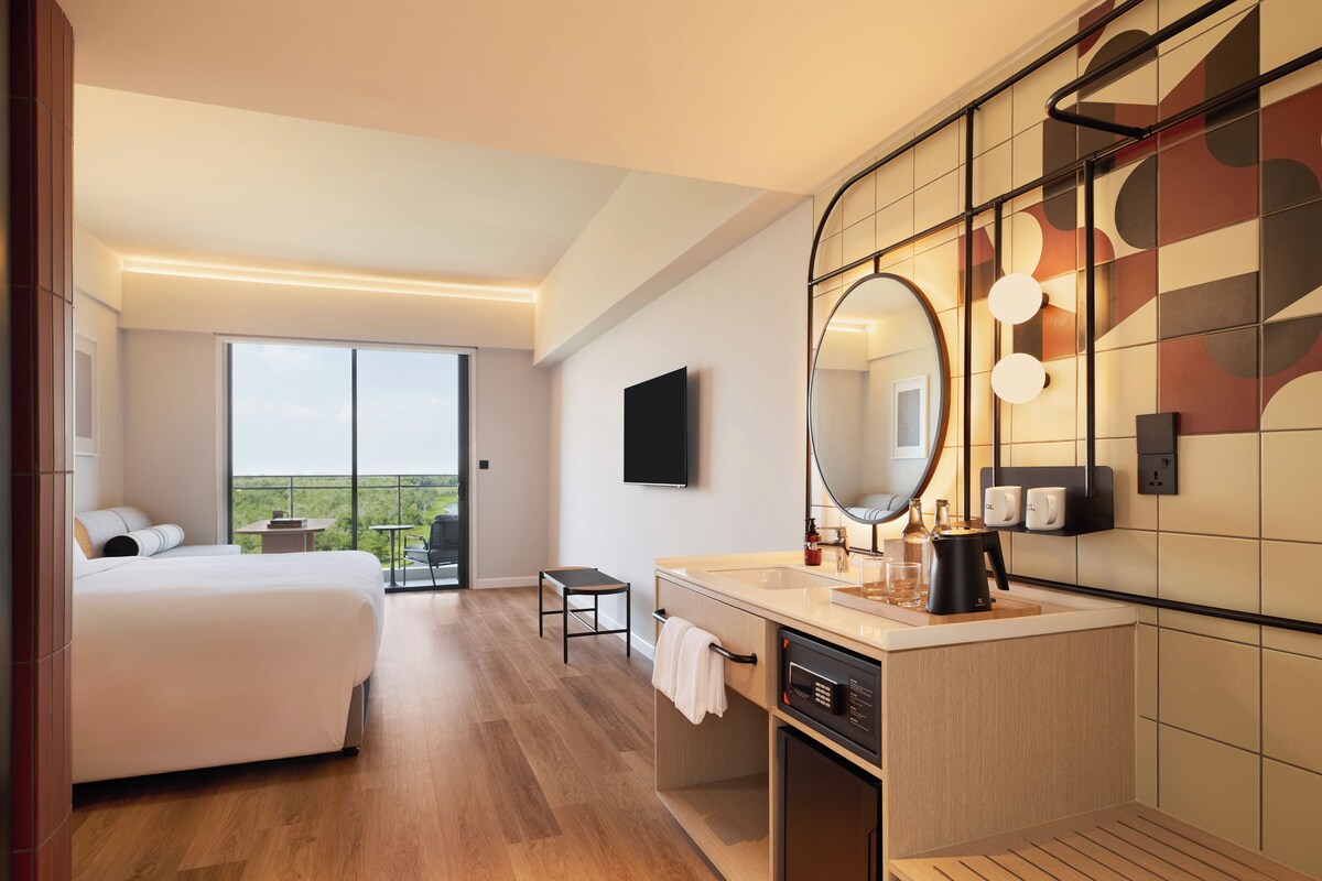 A hotel room with a bed, a desk, a chair, a large mirror, a sink, and a view of the outdoors.