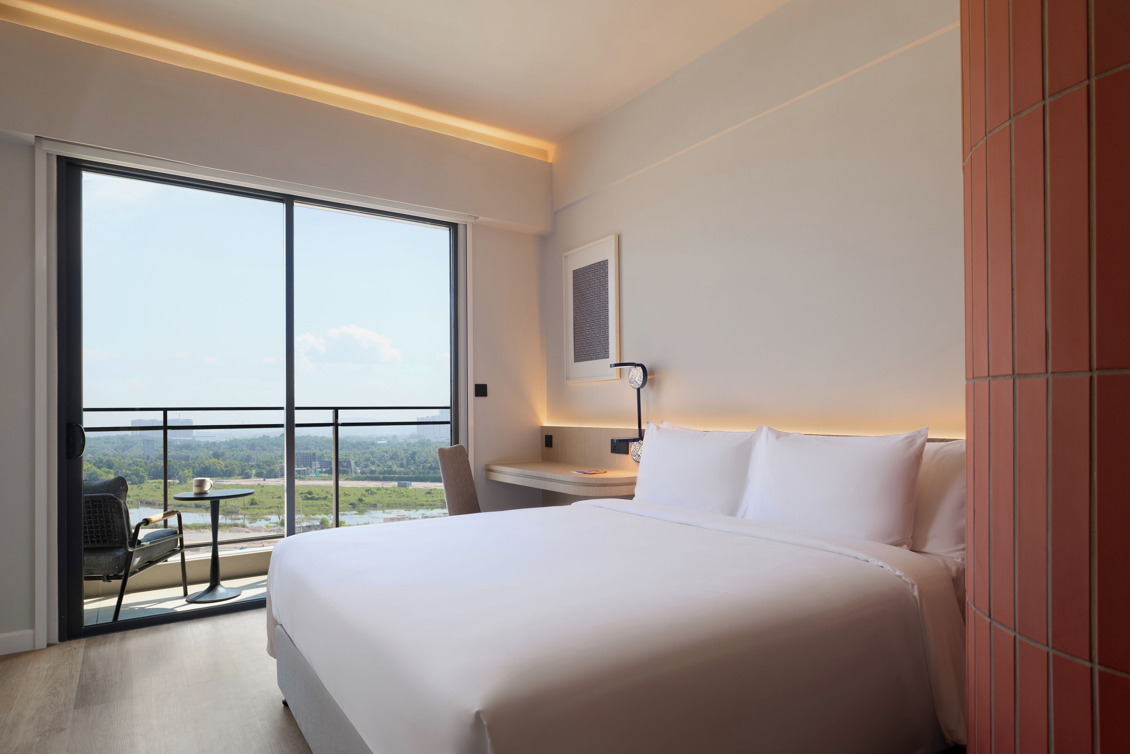 A hotel room with a large bed, a desk, a chair, and a view of a cityscape through a large window.