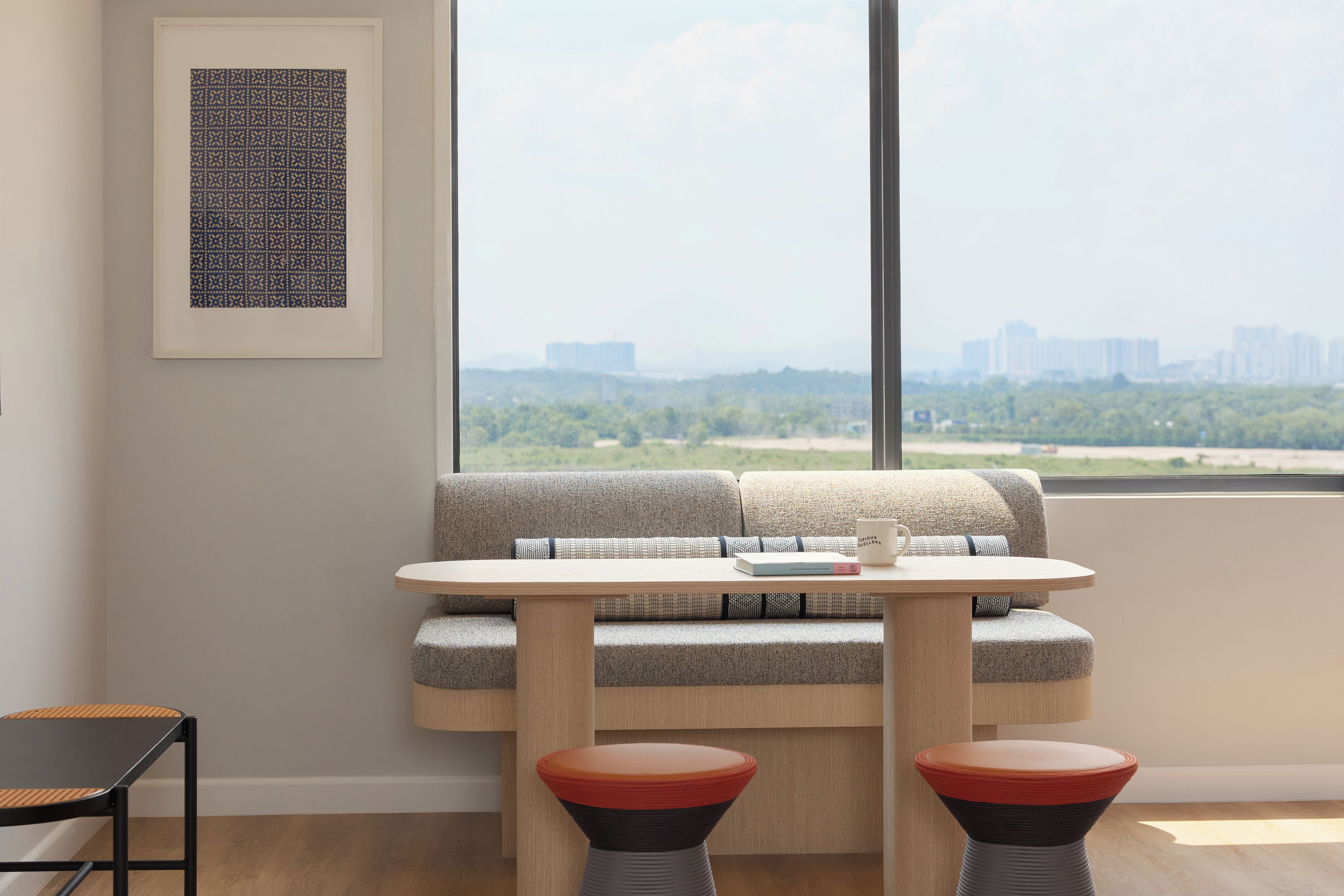 Una sala de estar moderna con un sofá, una mesa y dos taburetes. Hay una obra de arte enmarcada en la pared y una vista panorámica de la ciudad a través de la ventana.