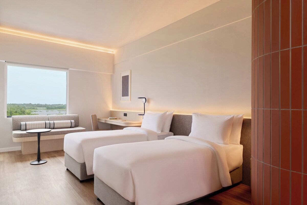A modern hotel room with two beds, a small table, a sofa, and a view of the outdoors.