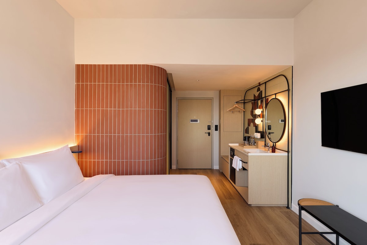 A hotel room with a bed, a bathroom with a mirror and sink, and a wooden floor.