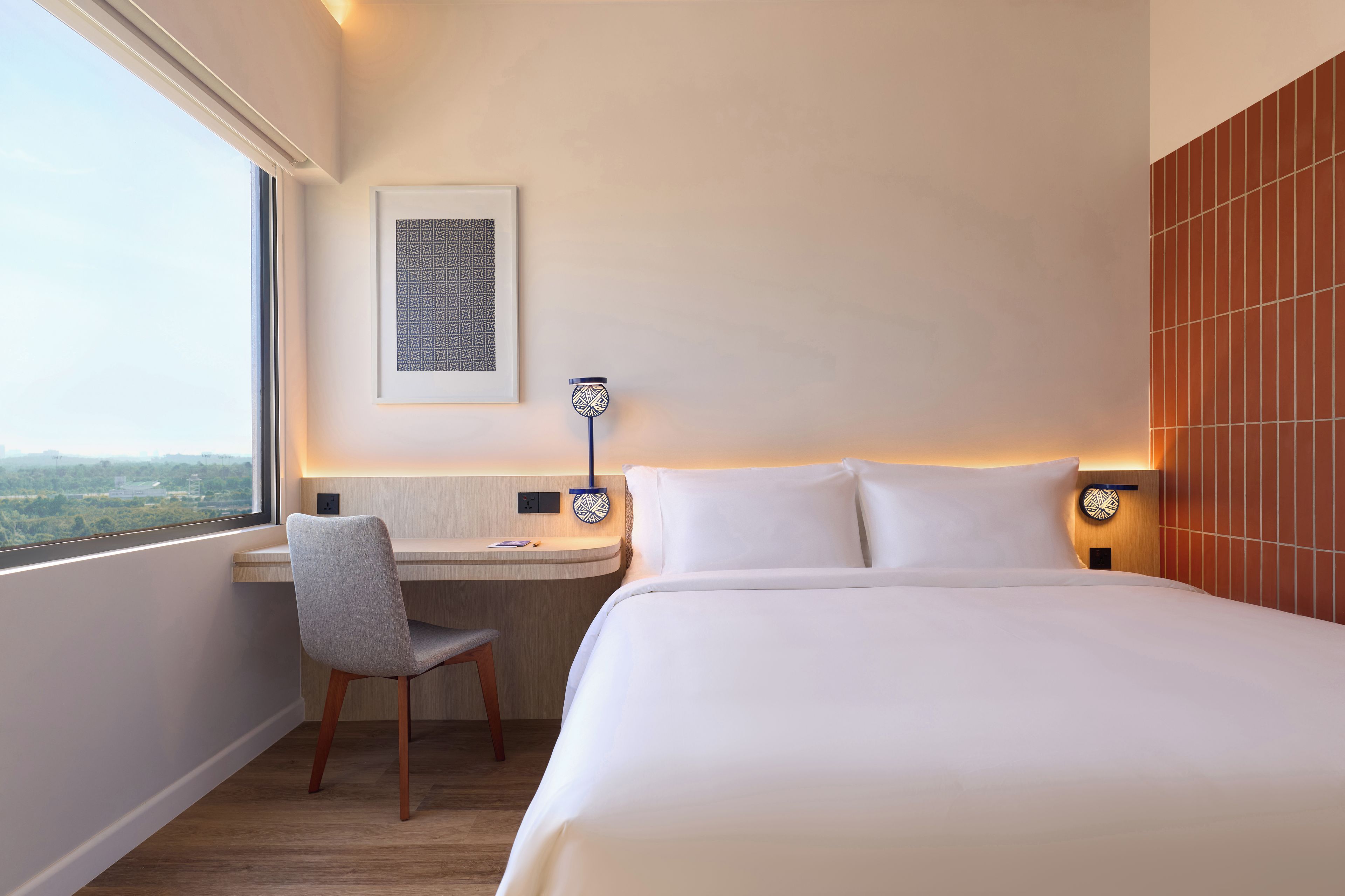 A hotel room with a bed, a desk, a chair, a window, and a wall-mounted lamp.