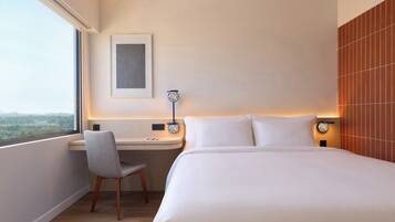 A hotel room with a bed, a desk, a chair, a window, and a wall-mounted lamp.
