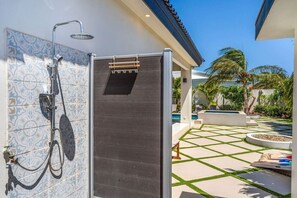 Zona de ducha exterior moderna con pared de azulejos, puerta de madera y vista a la piscina con palmeras y un diseño geométrico de azulejos.