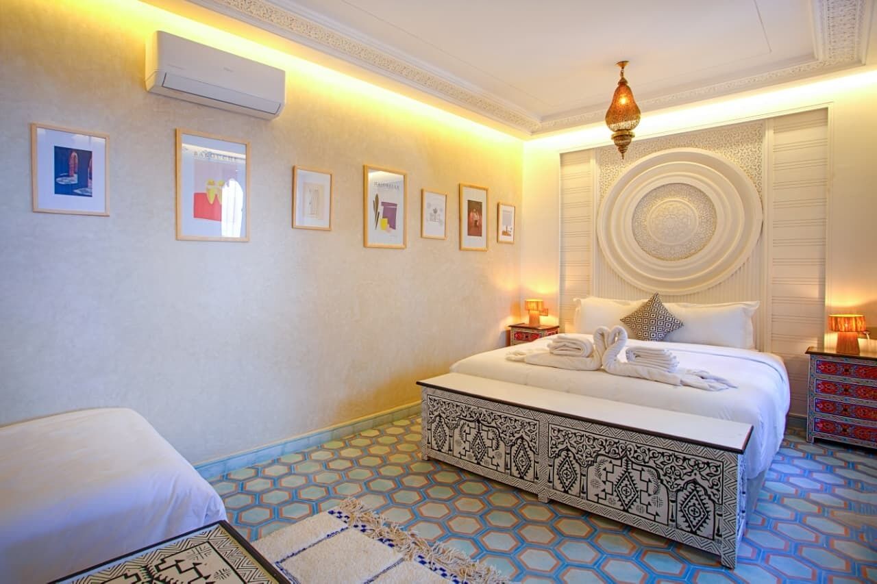 A hotel room with a bed, bedside table, wall-mounted air conditioning unit, framed pictures, decorative pillows, and a patterned floor.
