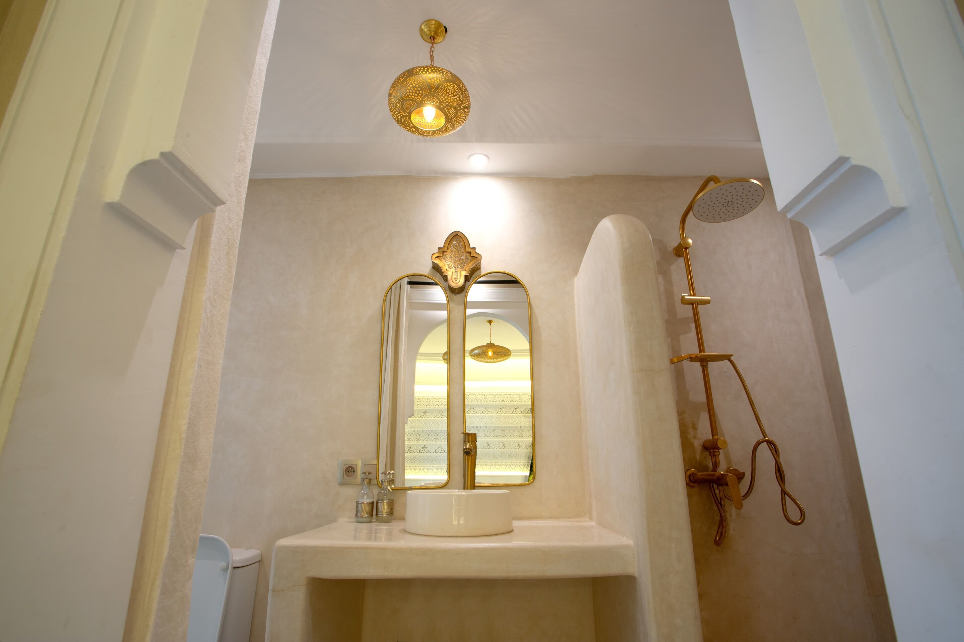 Een badkamer met een witte wastafel, een spiegel, een gouden douchekop en een hangende lamp.