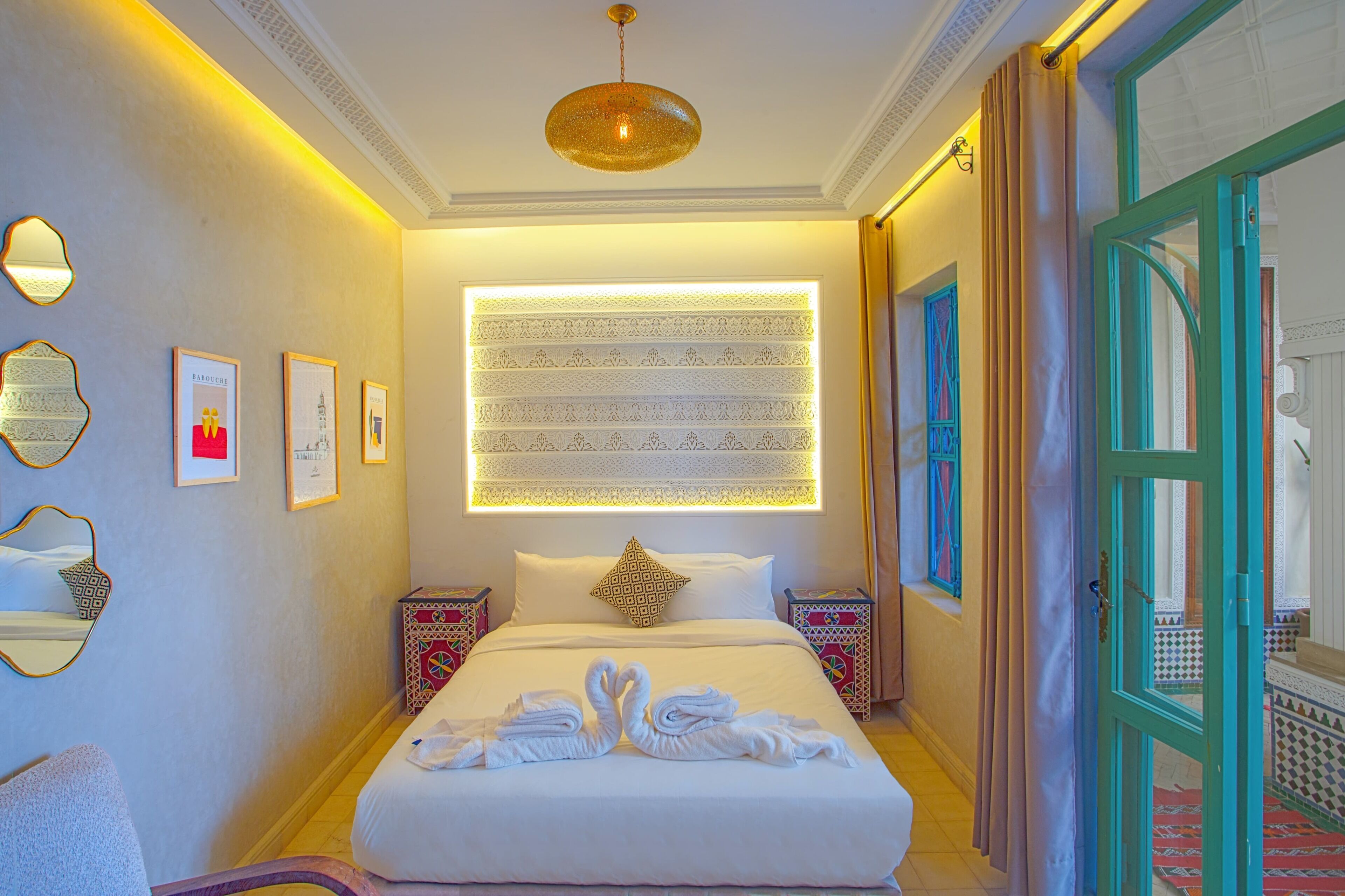 A hotel room with a large bed, decorative wall panels, a hanging light fixture, and a view through an open door.