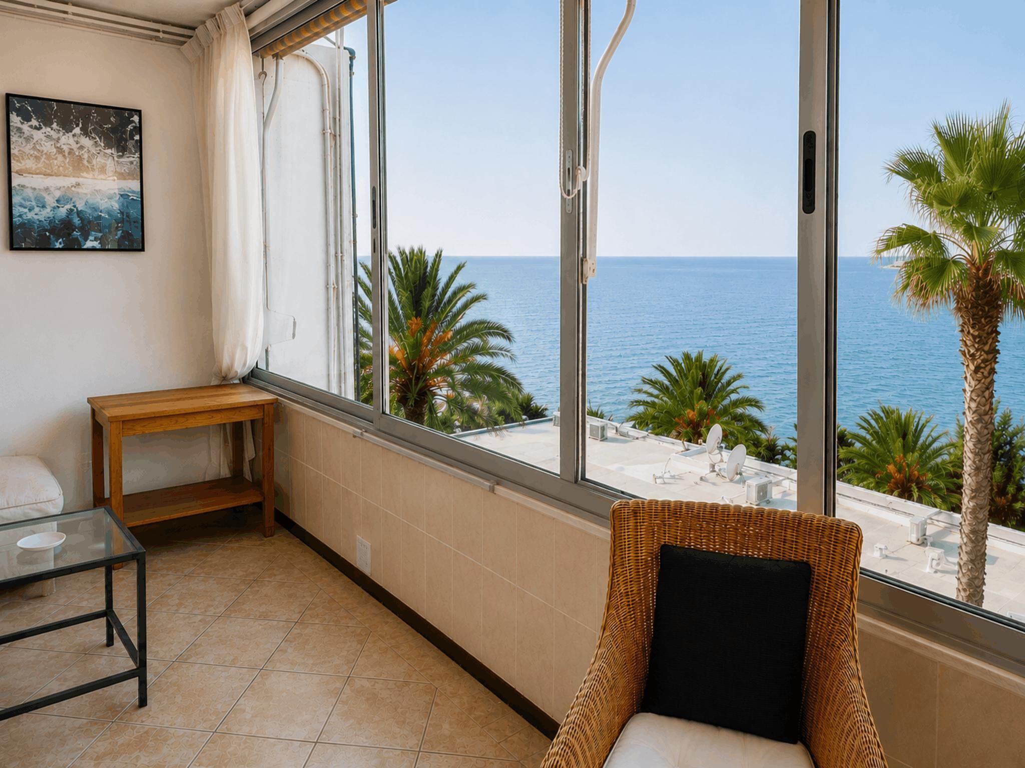 A balcony with a view of the sea, a wicker chair, a small table, and a painting on the wall.