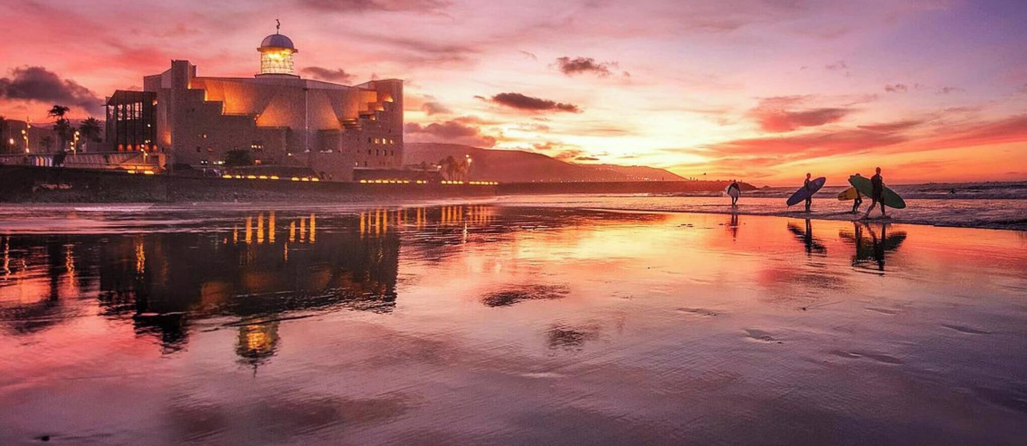 Atardecer sobre una playa con un edificio histórico y surfistas caminando.