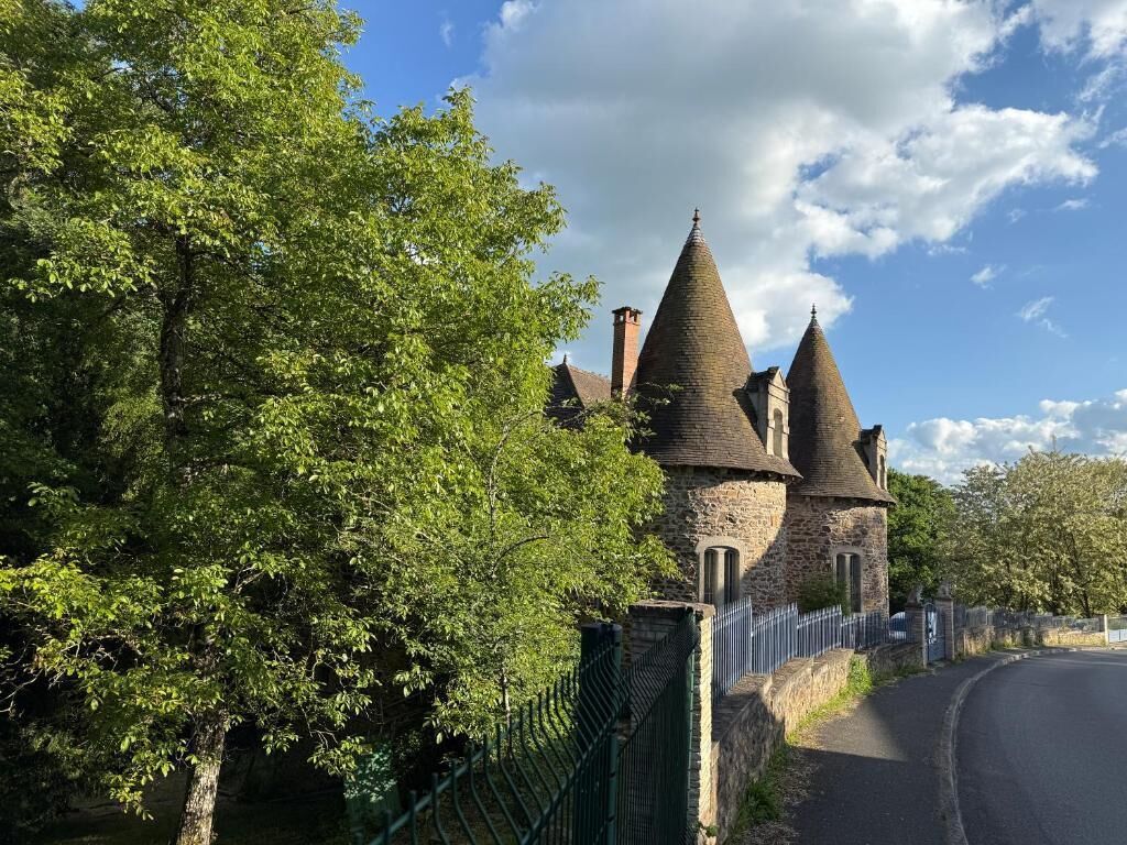 A stone building with multiple conical roofs, surrounded by trees and a metal fence.