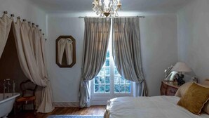A bedroom with a large bed, a chandelier, a window with curtains, a mirror, a bedside table with a lamp, and a wooden floor.