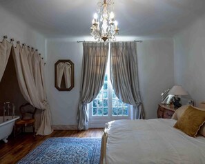 A bedroom with a large bed, a chandelier, a window with curtains, a mirror, a bedside table with a lamp, and a wooden floor.