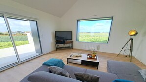 A modern living room with a grey sofa, a coffee table, and a TV. There are large windows offering a view of a green field. - House in Brittany with sea view for 2 people. (Cléder)