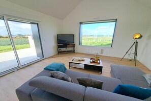 Ein modernes Wohnzimmer mit einem grauen Sofa, einem Couchtisch und einem Fernseher. Große Fenster bieten Blick auf ein grünes Feld.