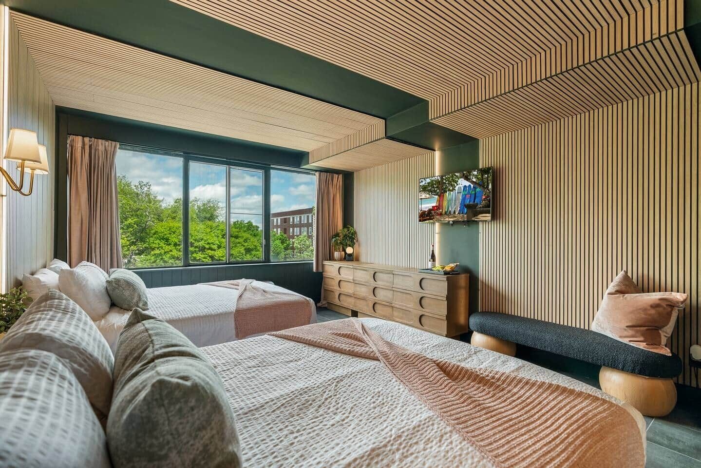 A modern bedroom with two beds, a dresser, a bench, and a large window offering a view of greenery.