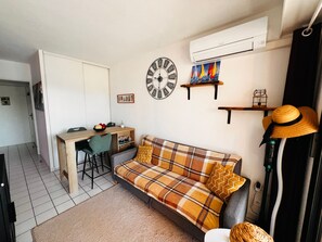 Ett kompakt vardagsrum med en soffa, ett litet matbord, en väggklocka och en luftkonditioneringsanläggning.