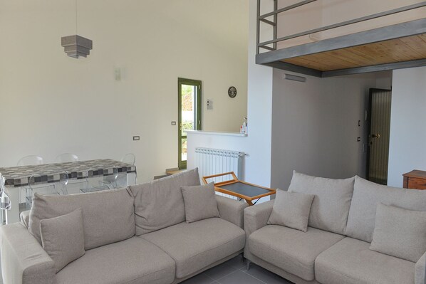 A modern living room with a sectional sofa, a dining table with chairs, and a kitchen area with a radiator.