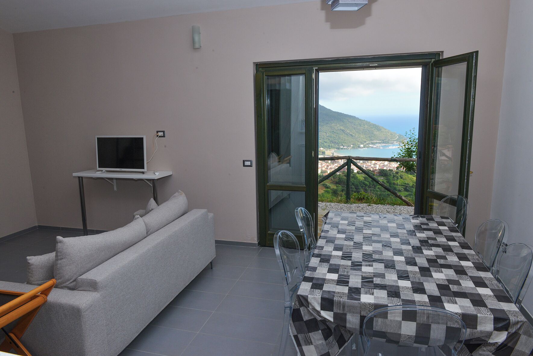 Ein Zimmer mit einer Couch, einem kleinen Fernseher auf einem Schrank, einem Tisch mit Stühlen und einem Balkon mit Blick.
