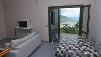 Ein Zimmer mit einer Couch, einem kleinen Fernseher auf einem Schrank, einem Tisch mit Stühlen und einem Balkon mit Blick.