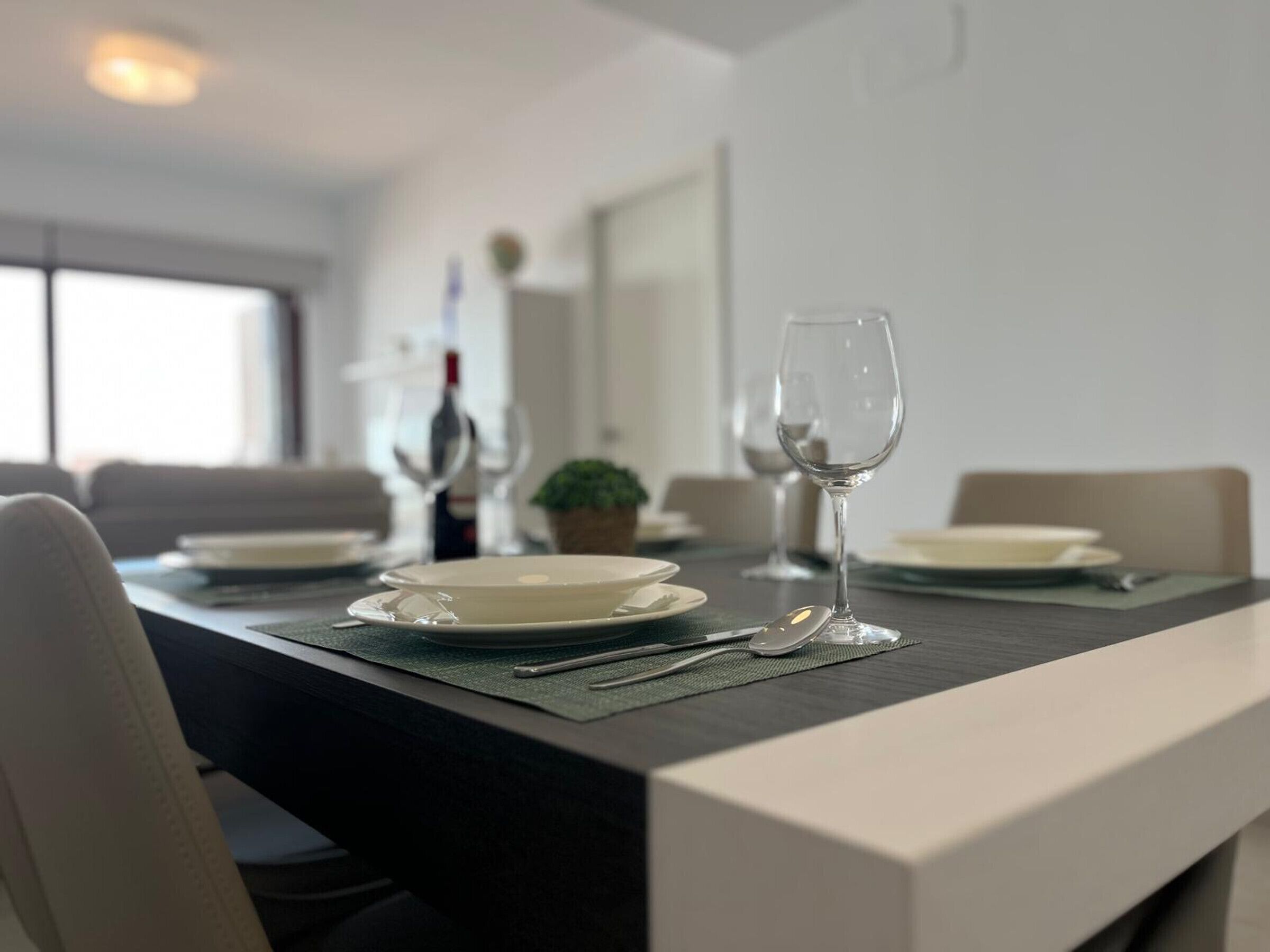 Mesa de comedor dispuesta con platos, cubiertos y copas de vino, con una botella de vino y una planta pequeña al fondo.