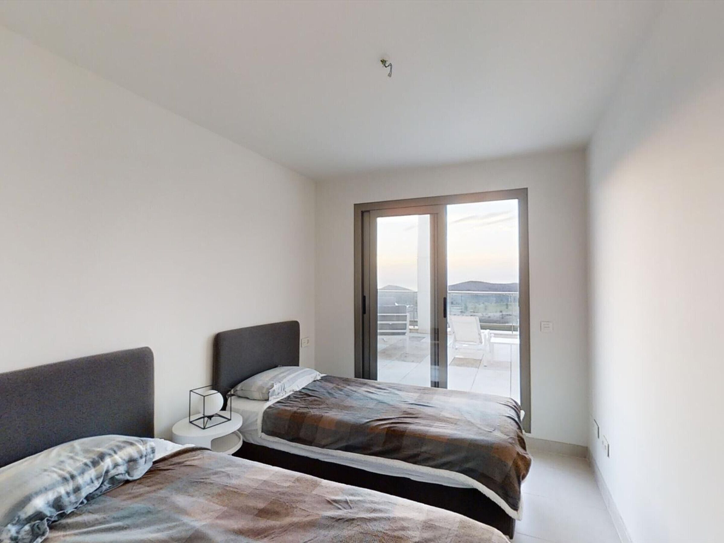 A modern bedroom with two beds, a nightstand, and a sliding glass door leading to an outdoor balcony.