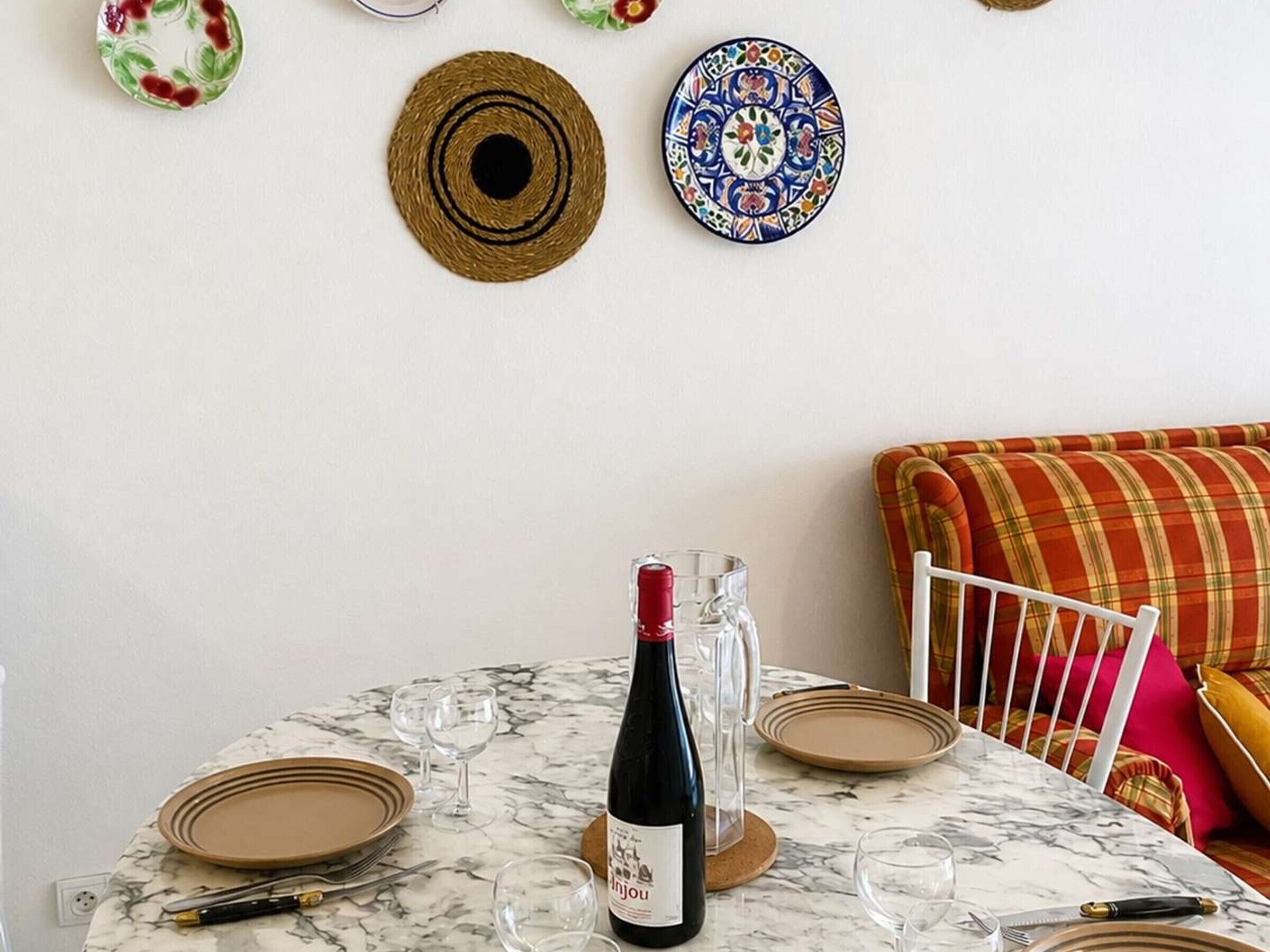 A dining table with plates, cutlery, and a wine bottle, set against a white wall with decorative plates and a plaid sofa.