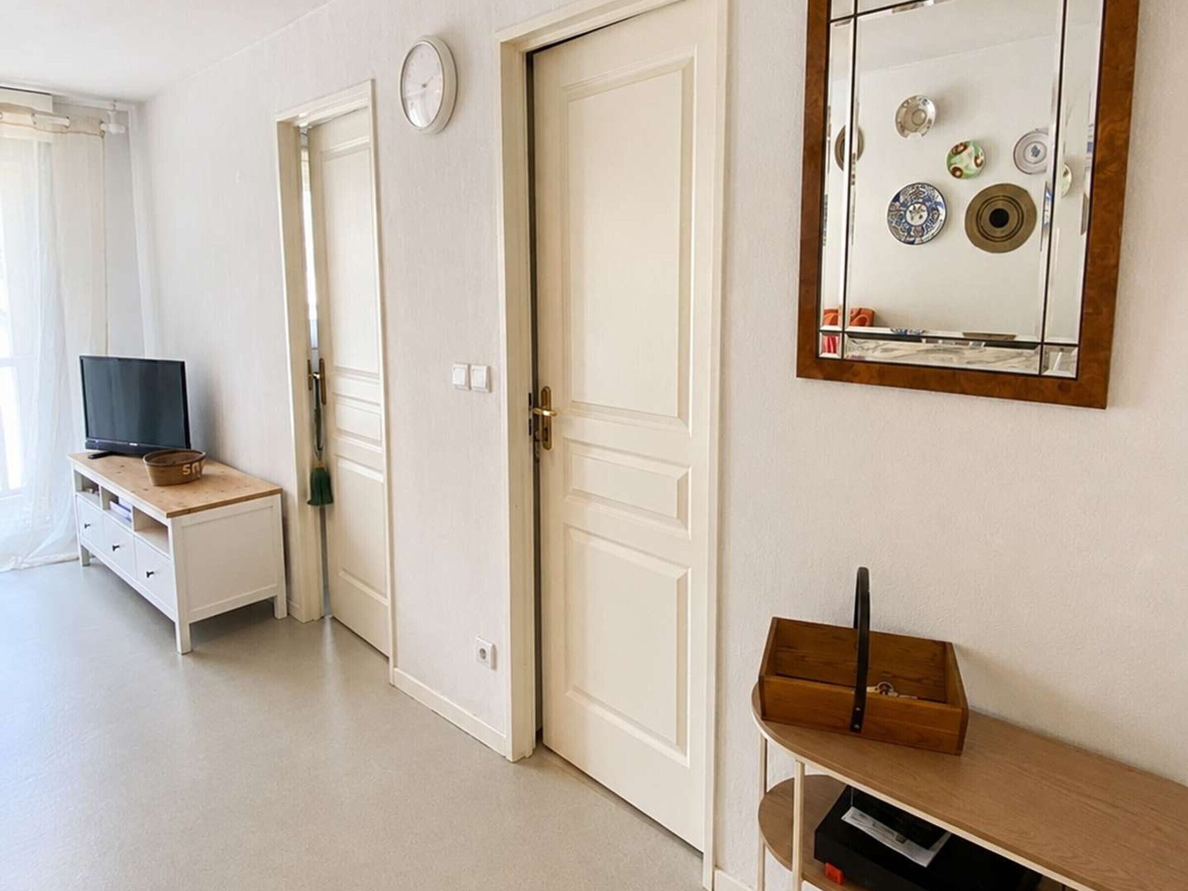 A room with a television on a wooden stand, a clock on the wall, and a mirror reflecting various items.