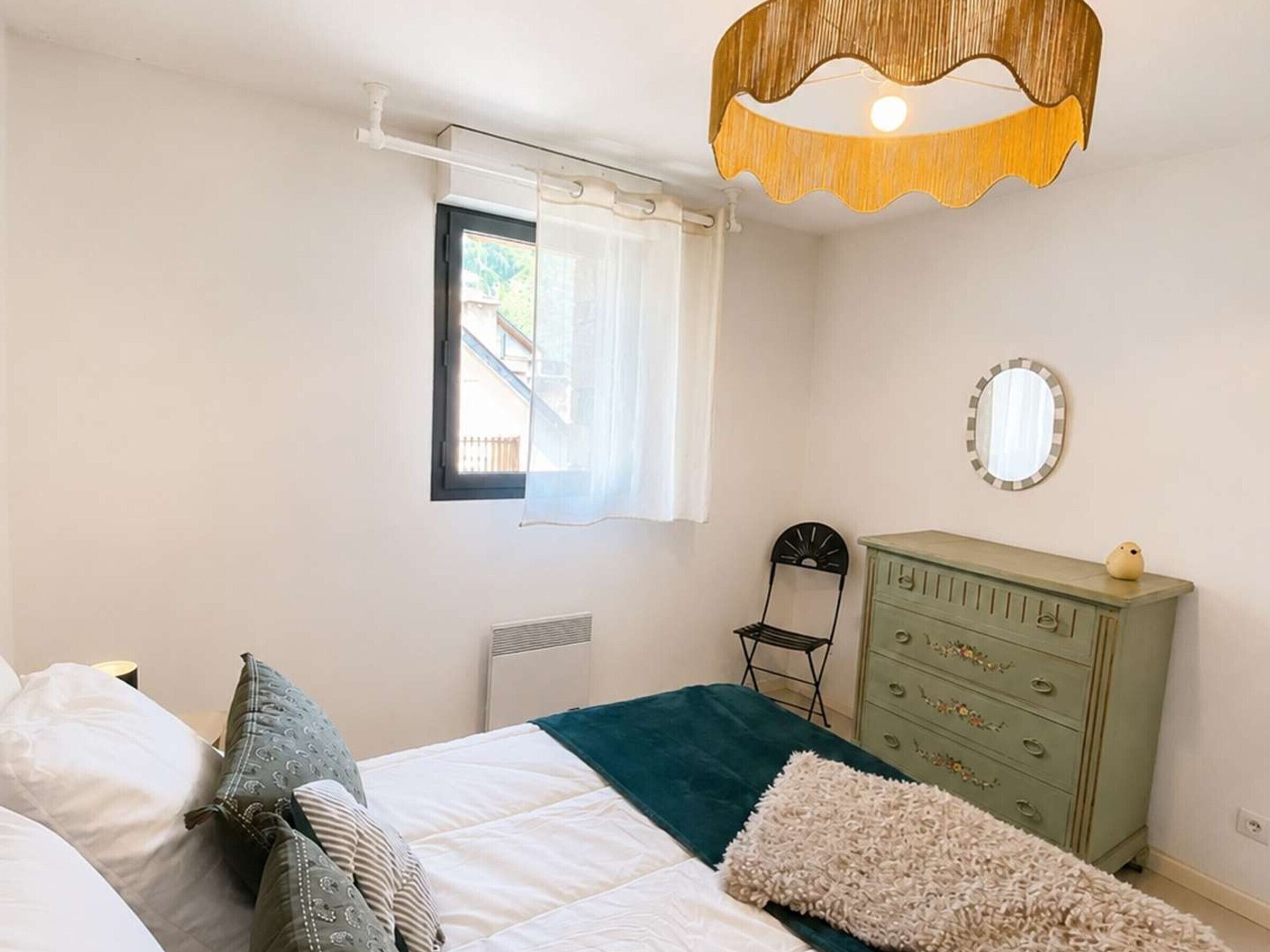 A bedroom with a bed, a dresser, a chair, a window with curtains, and a mirror on the wall.