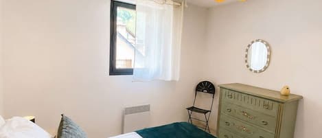 A bedroom with a bed, a dresser, a chair, a window with curtains, and a mirror on the wall.