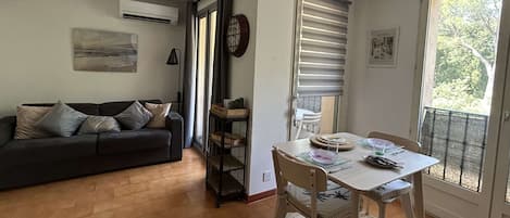 A living room with a sofa, a dining table with chairs, a small side table, and a window with blinds.