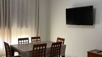 Een eethoek met een tafel, stoelen, een aan de muur bevestigde televisie en een raam met gordijnen.