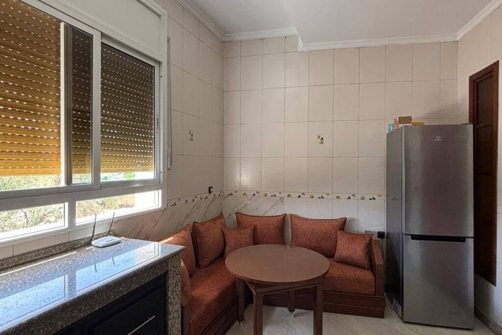 Una cocina con horno empotrado, refrigerador, ventana con persianas y una mesa redonda con sofá.