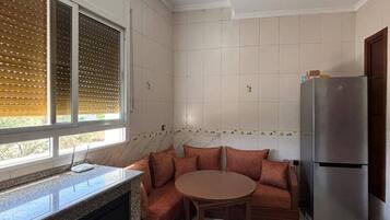 Una cocina con horno empotrado, refrigerador, ventana con persianas y una mesa redonda con sofá.