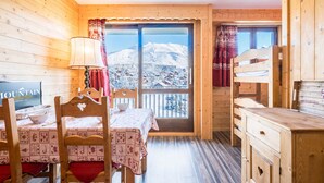 Quarto acolhedor com decoração em madeira, uma mesa de jantar com cadeiras, uma televisão e vista para montanhas cobertas de neve por uma grande janela.