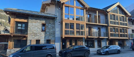 Un edificio de varios pisos con fachada de piedra y balcones de madera, que cuenta con una tienda a nivel de calle y coches estacionados.