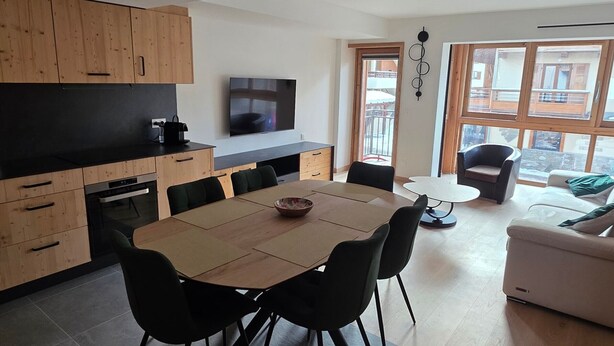 A modern kitchen with wooden cabinets, a dining area with a table and chairs, a TV mounted on the wall, and large windows offering a view of the outside. - Pleasant apartment, west balcony - 6 guests (Lanslevillard)